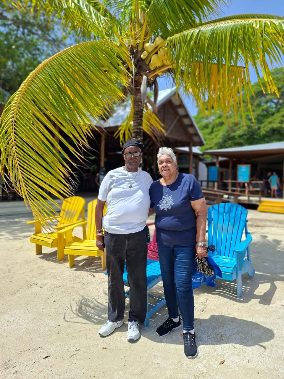 Carol and a travel companion standing under a palm tree beside bright beach chairs